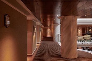 A hotel corridor with a marble column, carpeted floor, and a view of a lobby area.