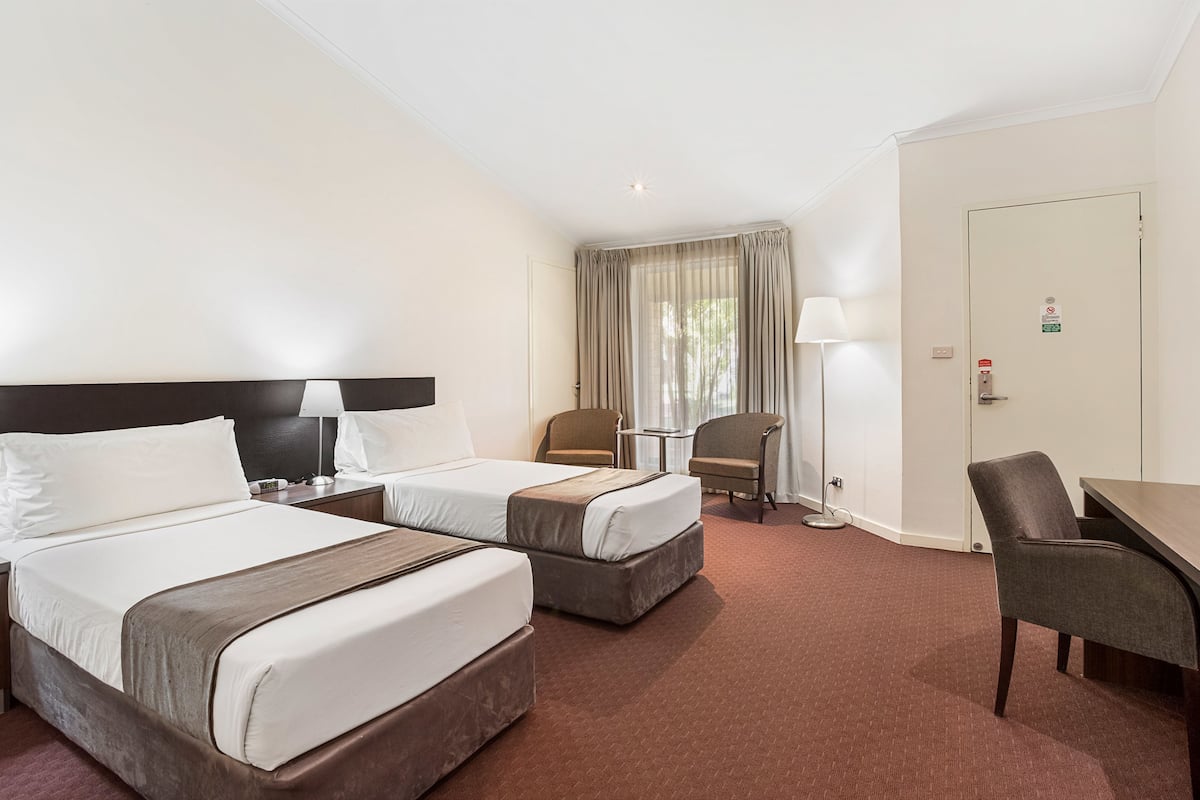 A hotel room with two beds, a desk, a chair, a lamp, and a window with curtains.