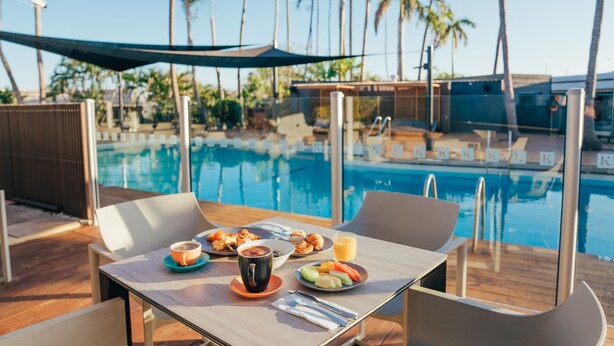 Une table à manger dressée avec des aliments pour le petit-déjeuner, donnant sur une piscine bordée de palmiers.