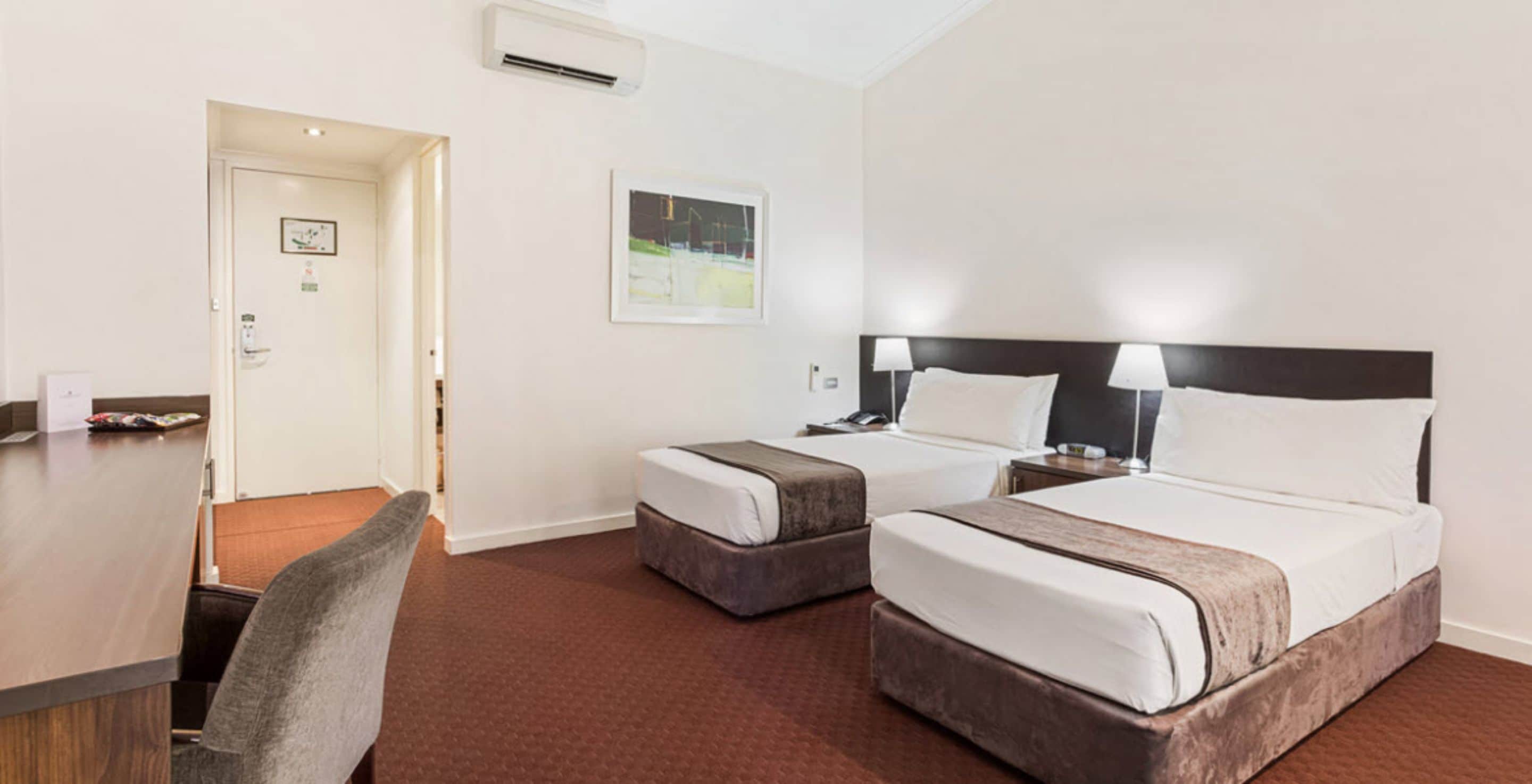 A hotel room with two beds, a desk, a chair, and a door.
