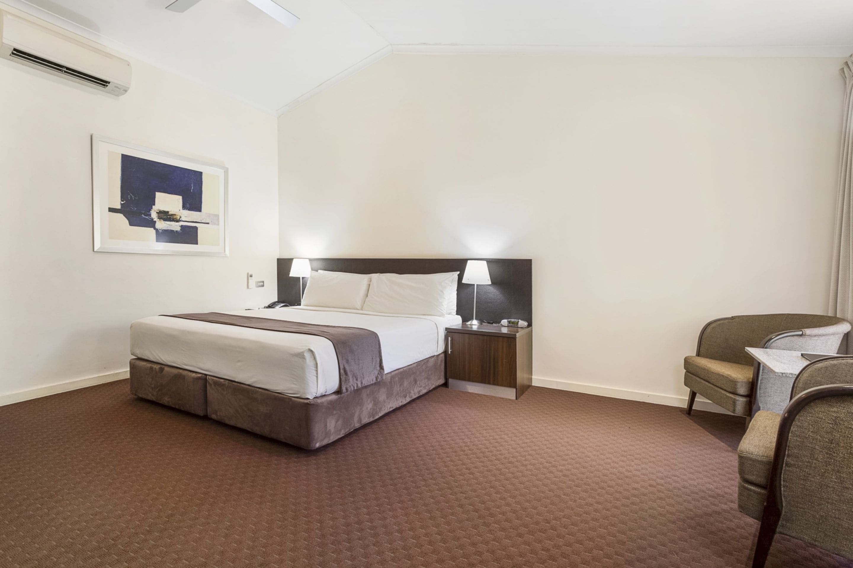 Una habitación de hotel con cama, mesitas de noche, una silla y un cuadro en la pared.