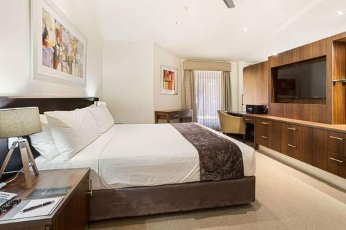 A hotel room with a bed, bedside table, lamp, and a TV mounted on the wall.