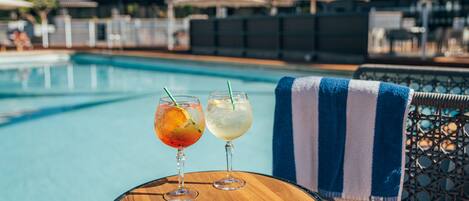 Une scène au bord de la piscine, avec deux boissons sur une table en bois, une serviette bleue et blanche à rayures, et des parasols en arrière-plan.