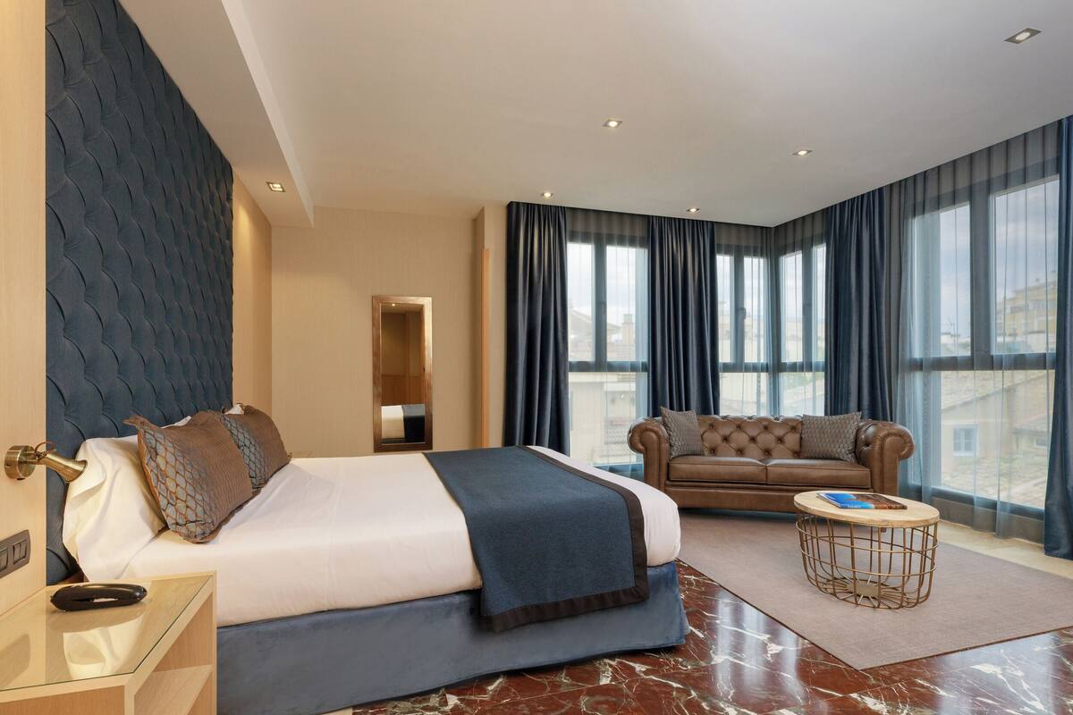 A modern hotel room with a large bed, a sofa, a round coffee table, and a view of the city through large windows.