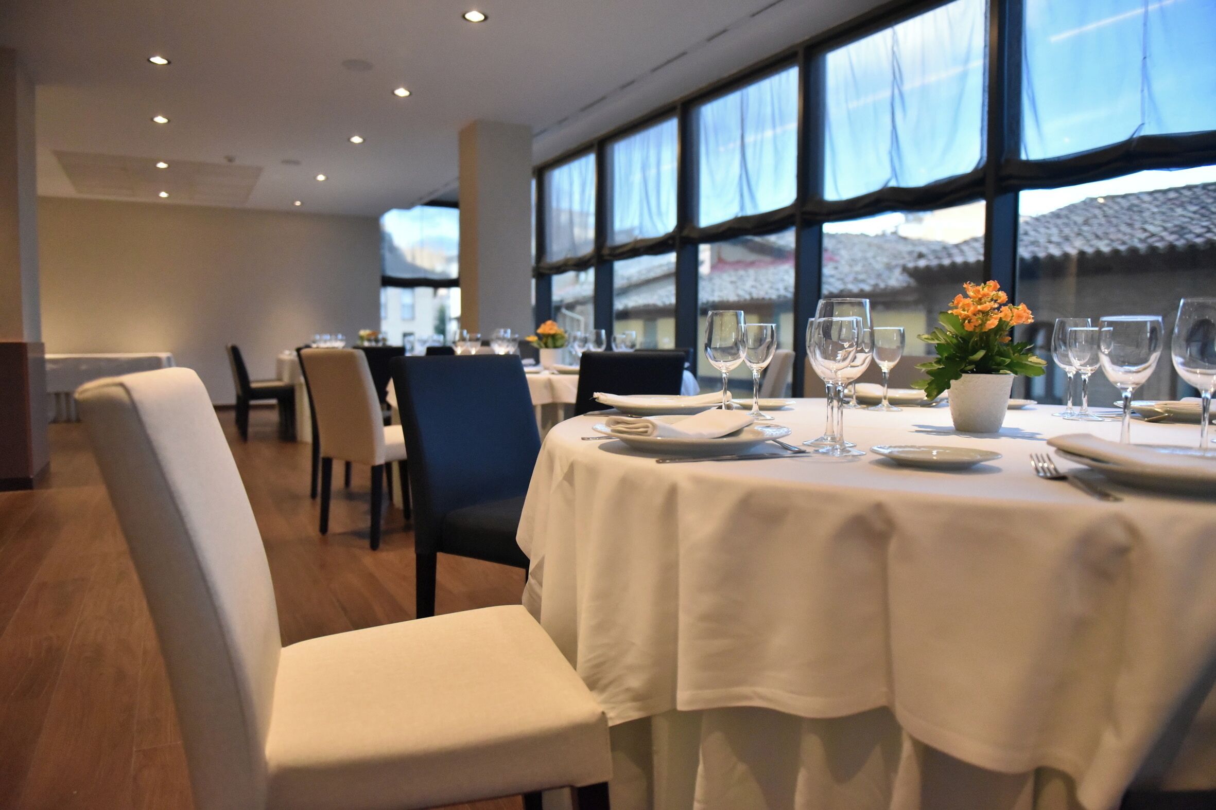 Un restaurant amb taules rodones preparades per dinar, grans finestres i vista d'un edifici.