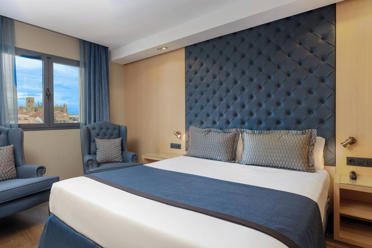 A modern hotel room with a large bed, a tufted headboard, a seating area with a chair and a sofa, and a view of the city through the window.