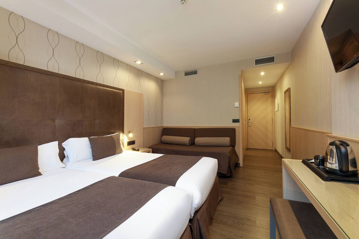 A hotel room with two beds, a desk with a kettle, and a TV mounted on the wall.