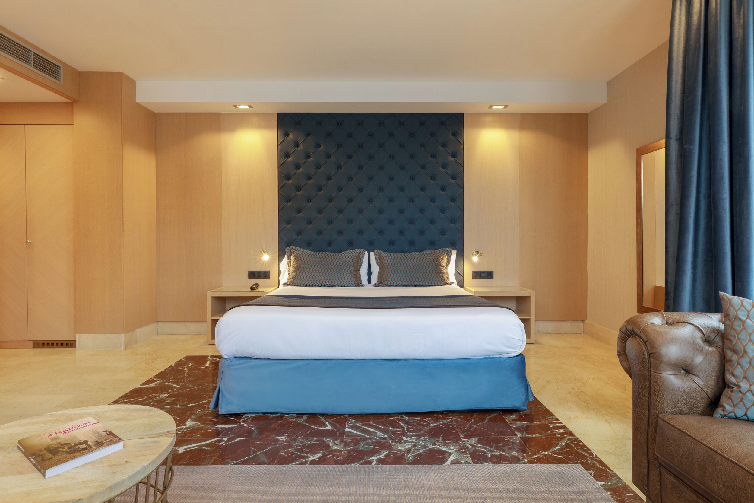 Una habitación de hotel moderna con una cama grande, un sofá de cuero, un tapete con diseño de mármol y una mesita con un libro.