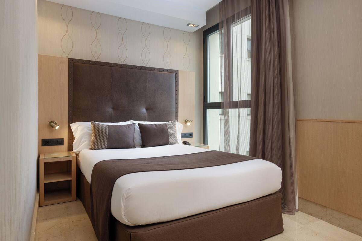 A modern hotel room with a large bed, a nightstand, a wall-mounted light, and a window with curtains.