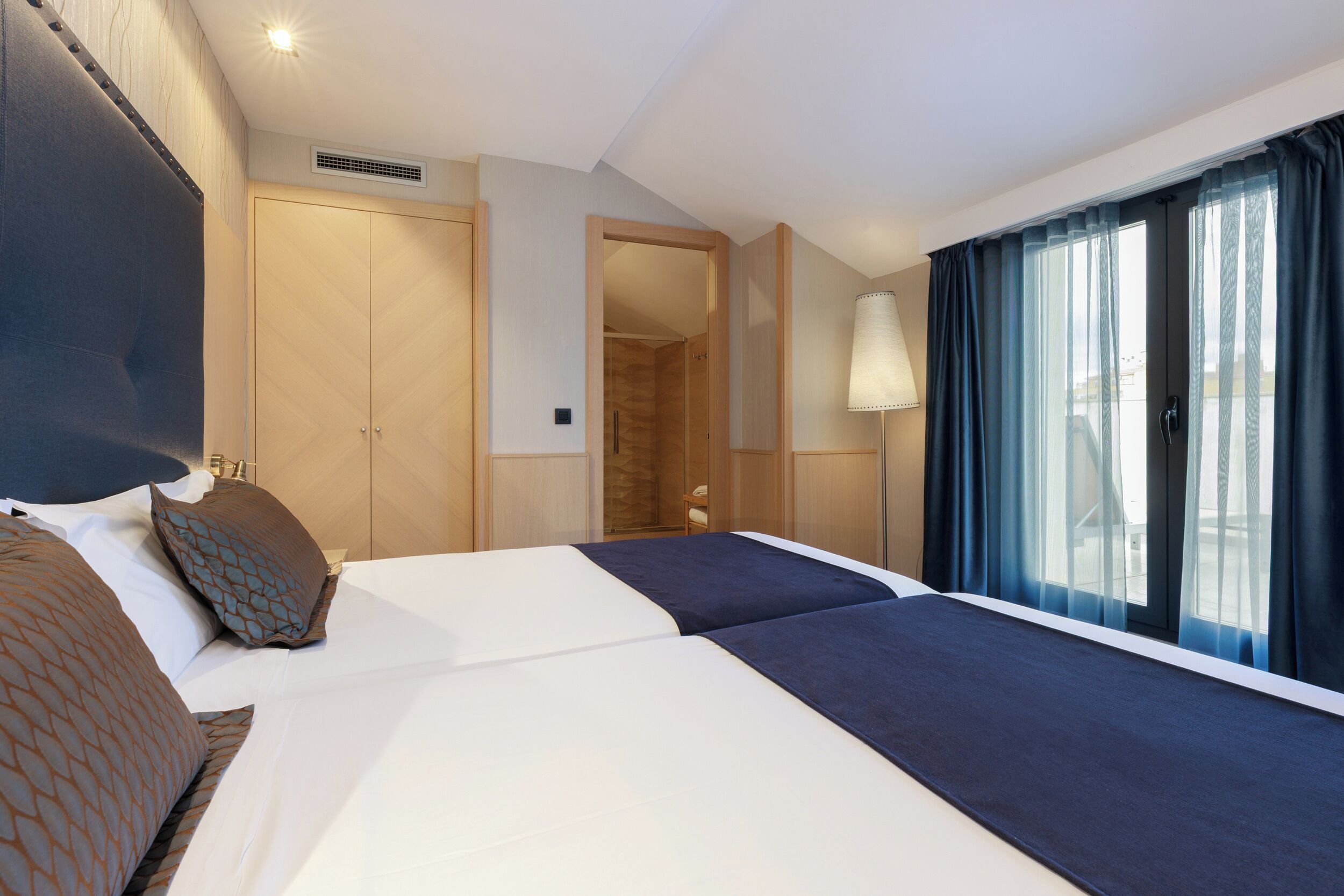 A hotel room with two beds, a large window with curtains, a wardrobe, and a lamp.