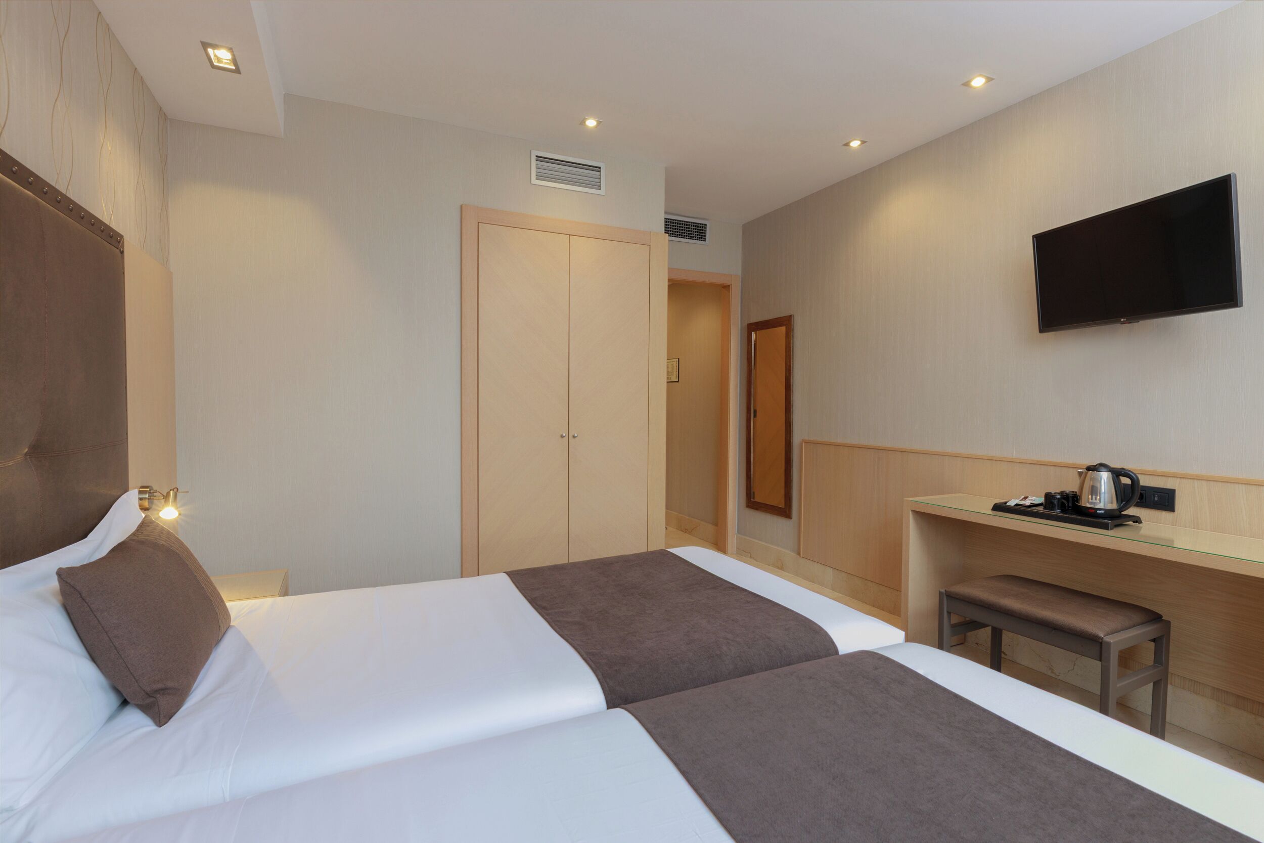 A hotel room with two beds, a TV, a desk with a kettle, and a wardrobe.
