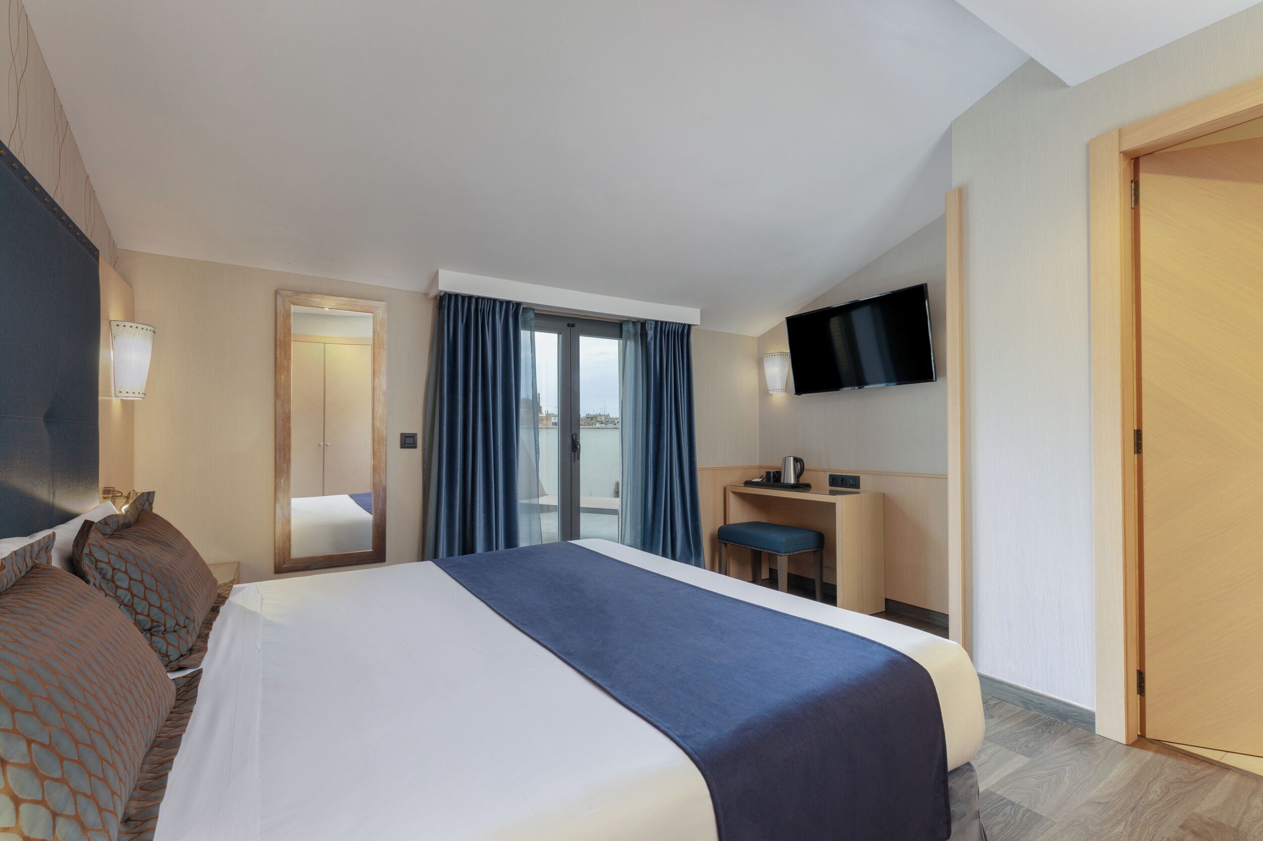 A hotel room with a large bed, a desk with a chair, a flat-screen TV, and a sliding glass door to the balcony.