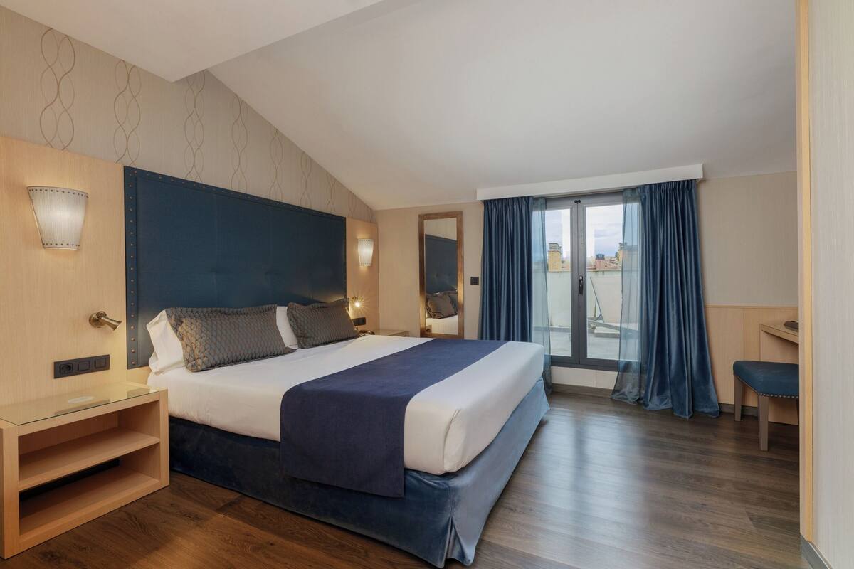 A modern hotel room with a large bed, a bedside table, a desk, and a view of the city through the window.