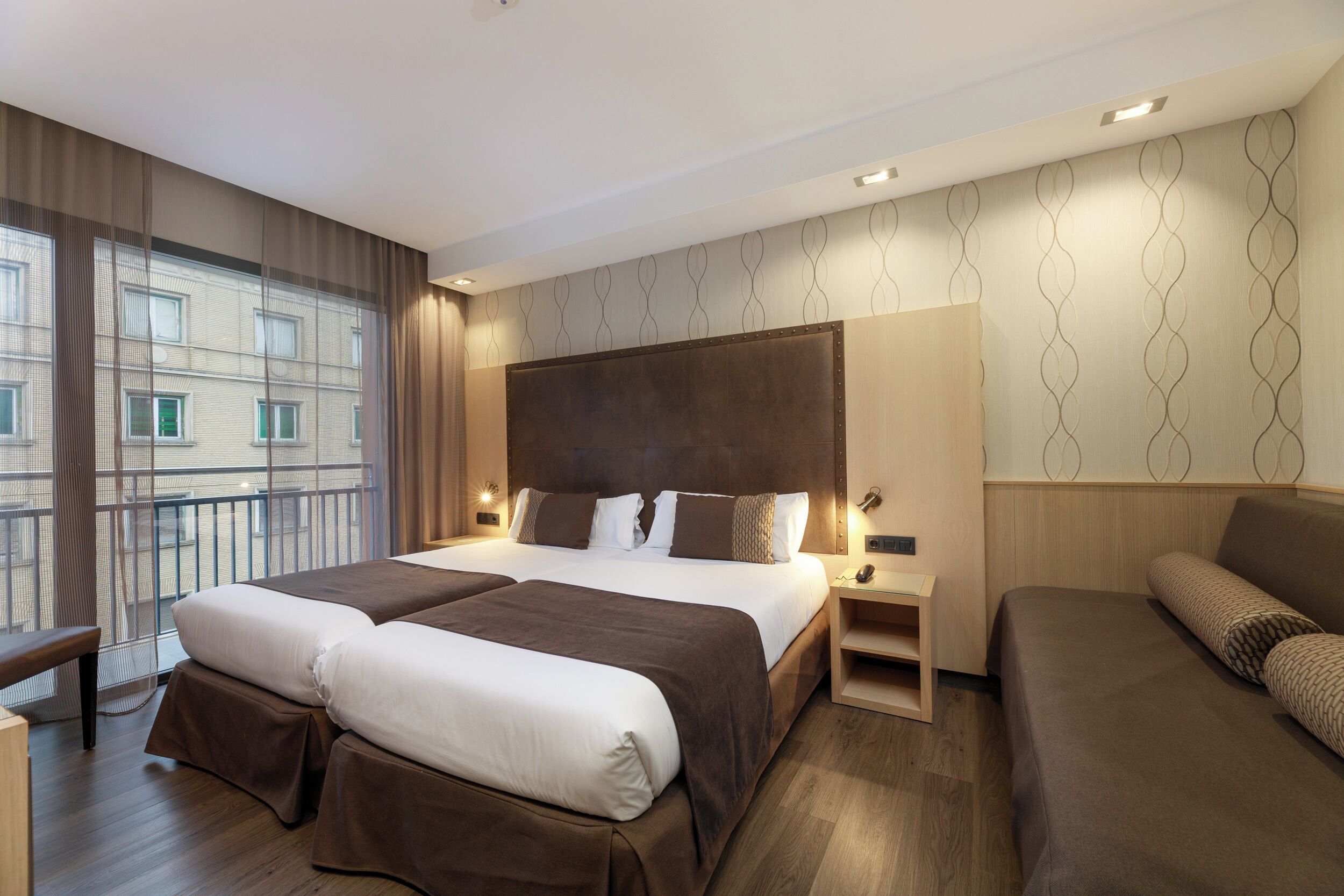 Una habitación de hotel moderna con una cama grande, dos mesitas de noche, un sofá y vistas a un paisaje urbano a través de amplias ventanas.