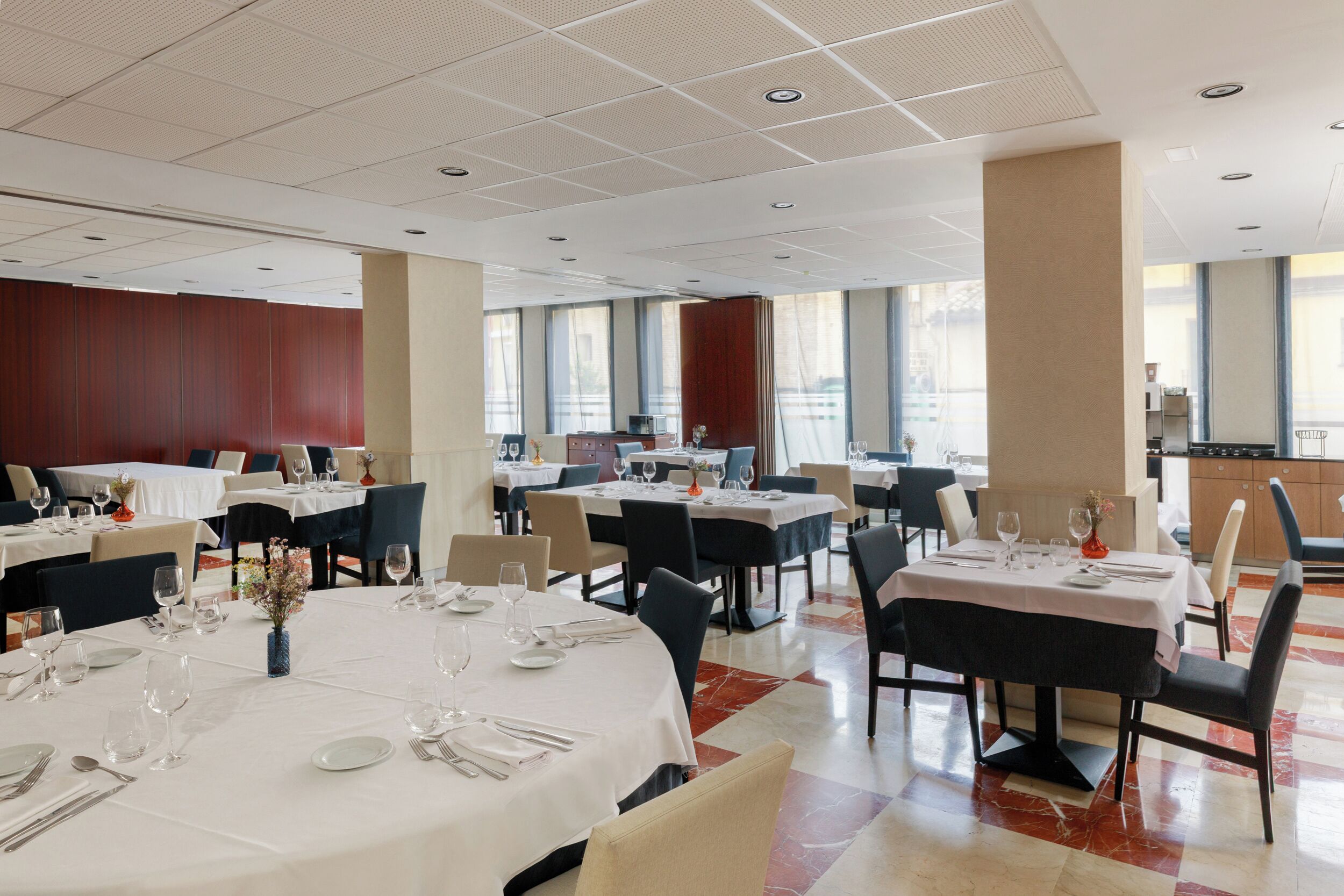A spacious hotel dining area with multiple tables set for dining, large windows offering views, and a modern ceiling design.