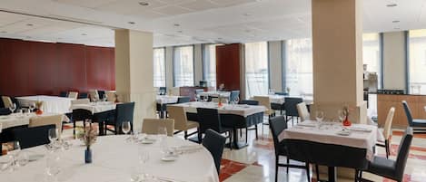 A spacious hotel dining area with multiple tables set for dining, large windows offering views, and a modern ceiling design.