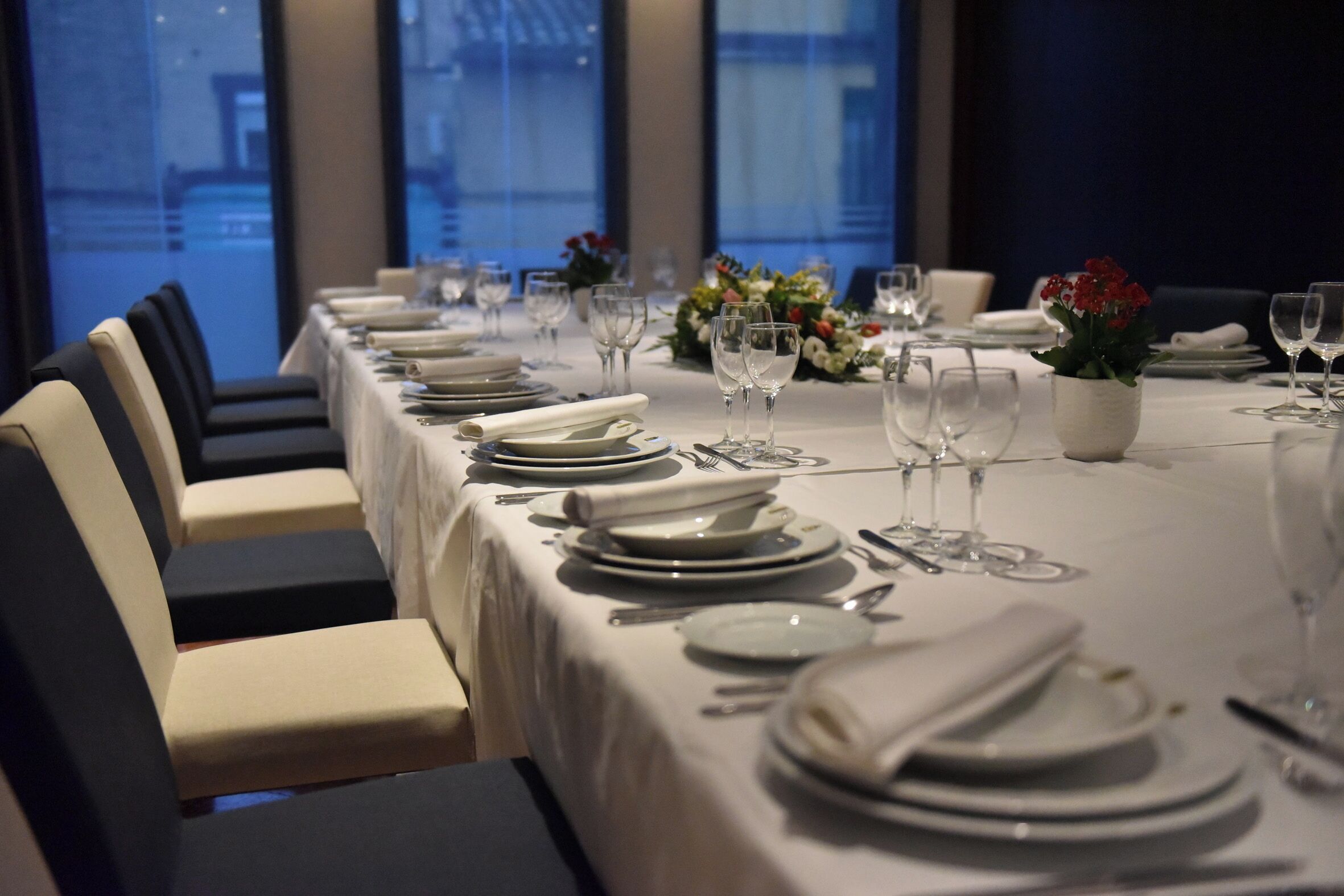 A dining table set with white tablecloth, plates, glasses, and cutlery, with chairs and a vase of flowers.