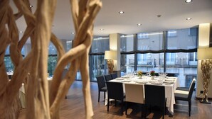 A modern dining area with a rectangular table set for six, surrounded by dark chairs. The room features large windows with a view of the outside, and the floor is wooden.