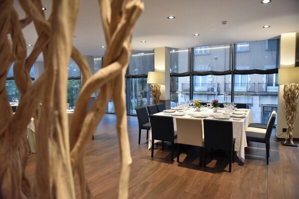 Un comedor moderno con una mesa rectangular dispuesta para seis personas, rodeada de sillas oscuras. La habitación tiene ventanas grandes con vistas al exterior, y el suelo es de madera.