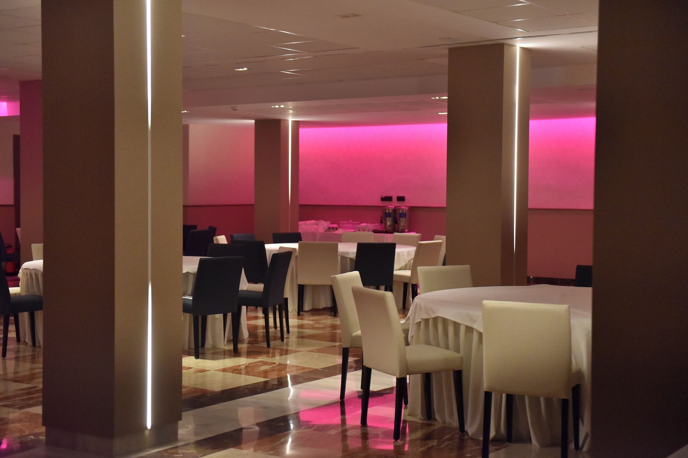 A spacious dining area with multiple tables and chairs, featuring a checkered floor and pink ambient lighting.