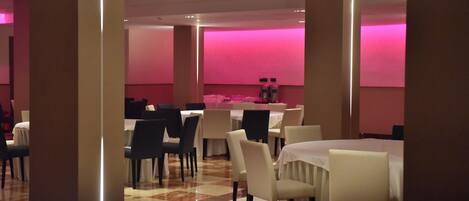 A spacious dining area with multiple tables and chairs, featuring a checkered floor and pink ambient lighting.
