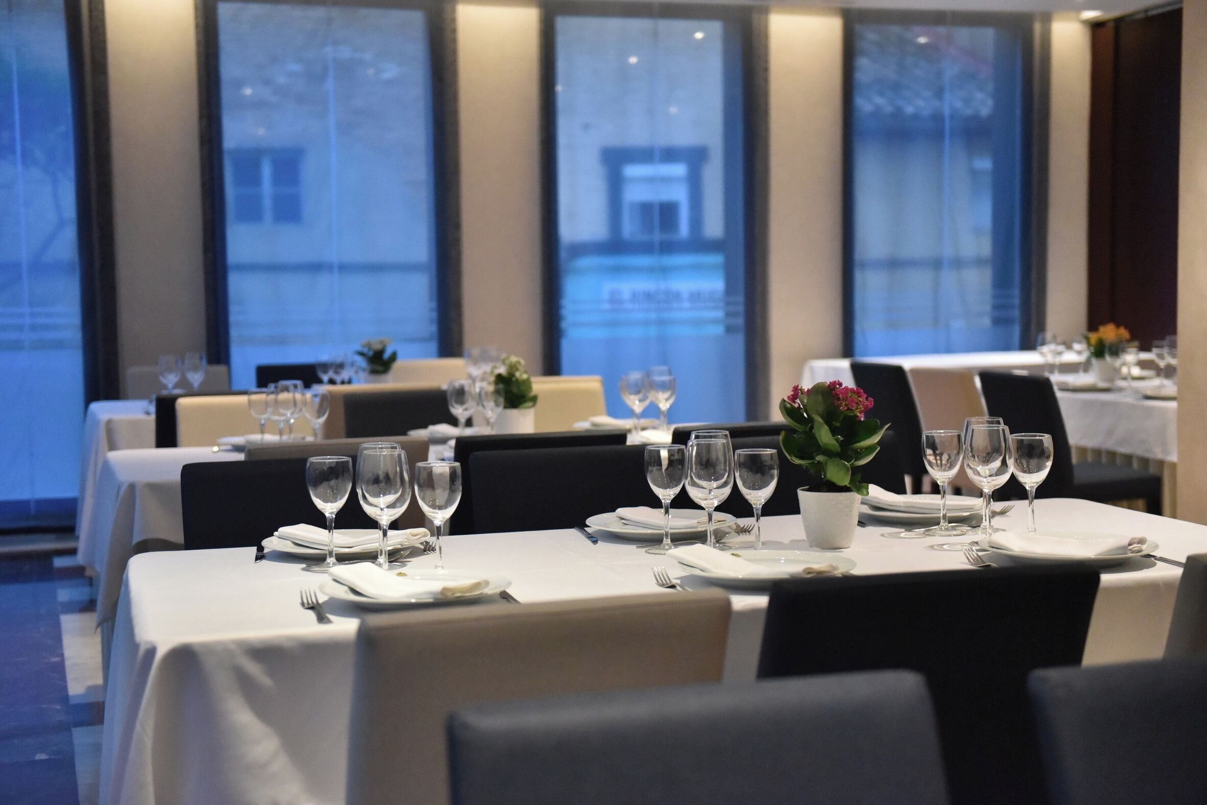 A dining area with tables set for a meal, featuring white tablecloths, wine glasses, and a potted plant.