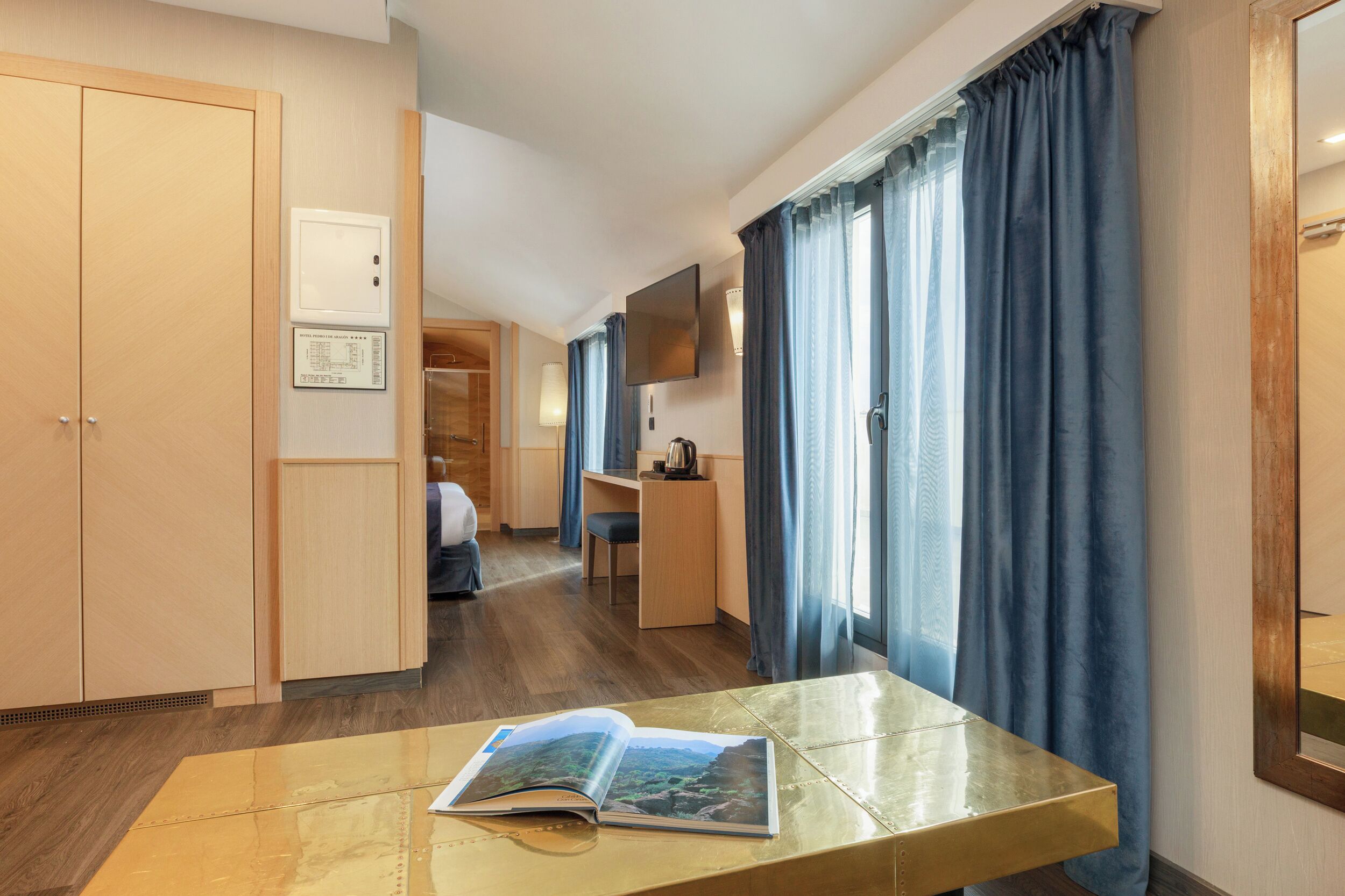 Habitación de hotel con un armario de madera, un escritorio con silla, un televisor de pantalla plana en la pared y una ventana con cortinas azules.