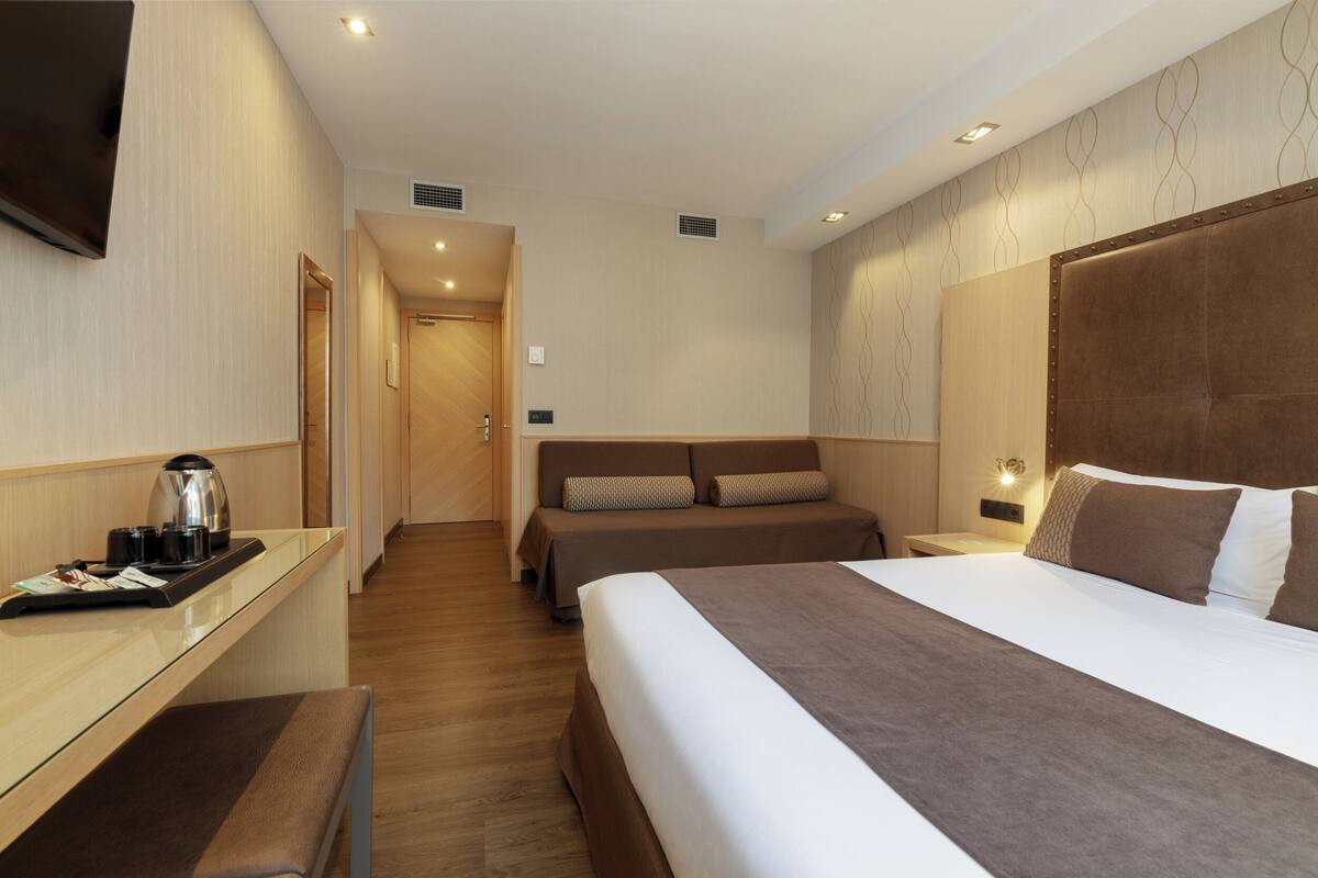 A hotel room with a large bed, a sofa, a desk with a kettle, and a TV on the wall.
