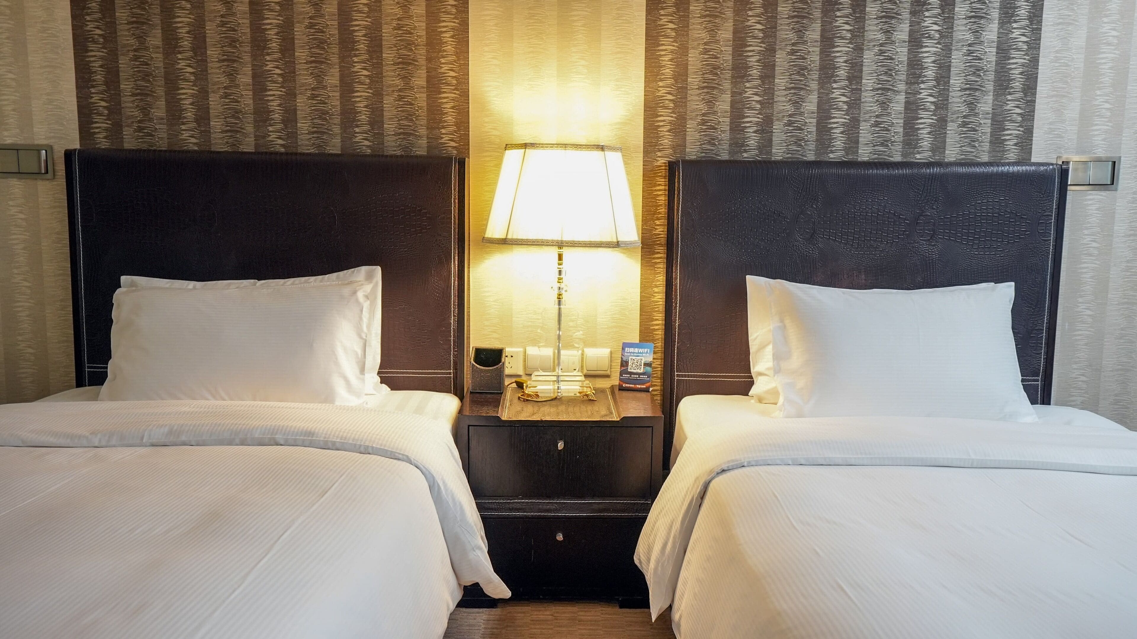 A hotel room with two beds, a nightstand with a lamp, and a wall-mounted outlet.
