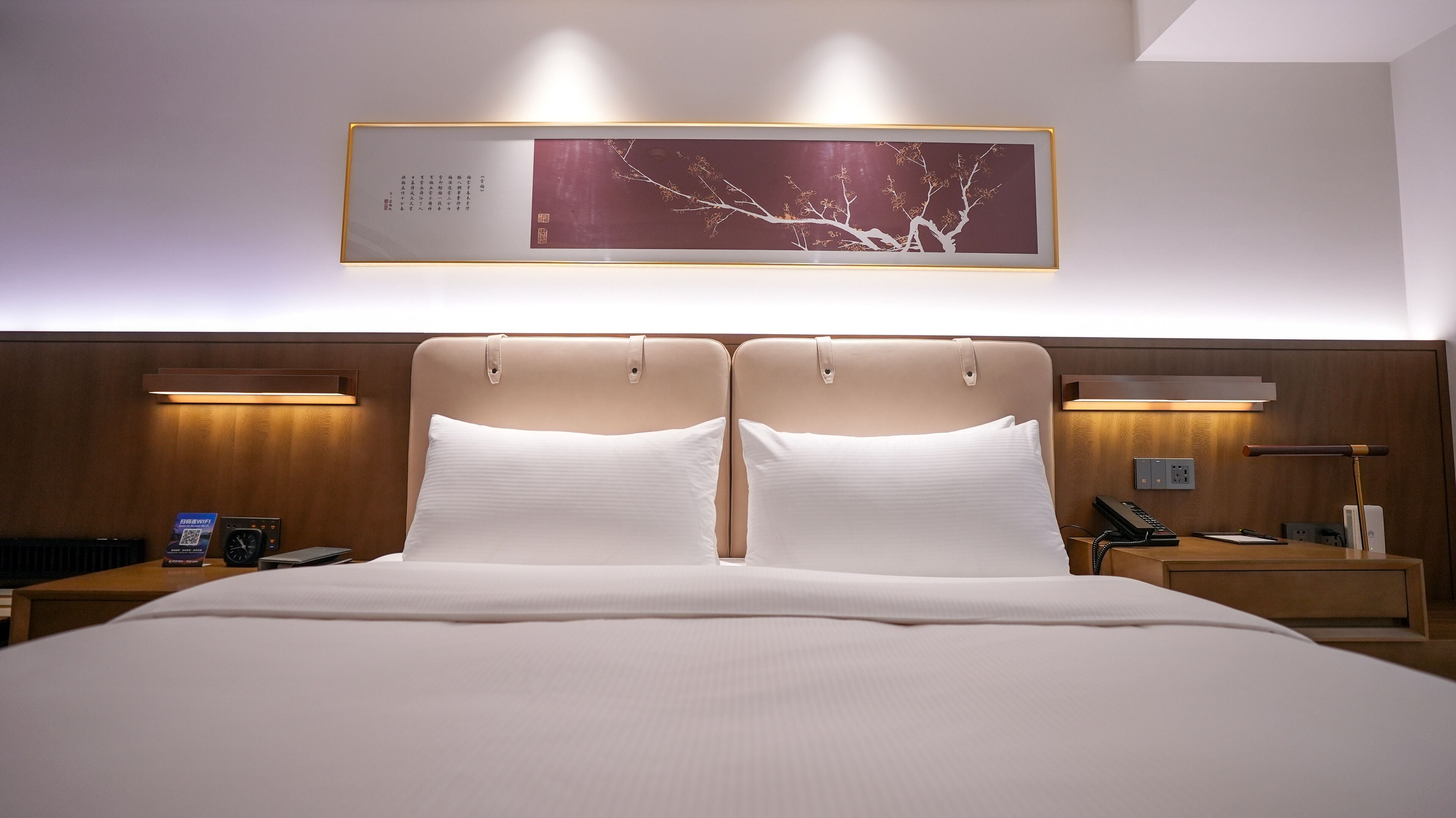 Quarto de hotel com uma cama grande, duas mesas de cabeceira, um criado-mudo, uma luminária na parede e uma obra de arte emoldurada sobre a cama.