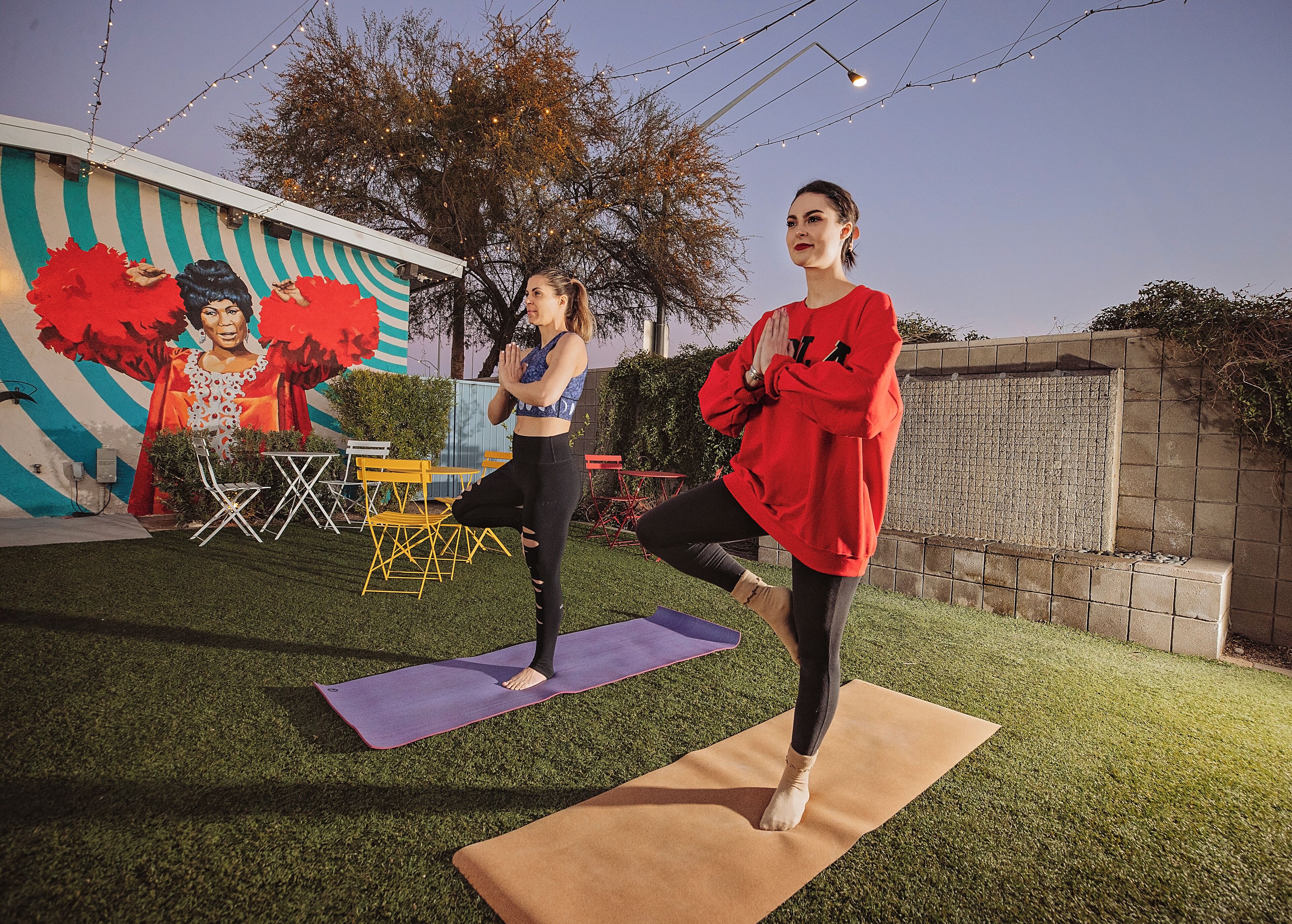 Zwei Personen machen Yoga im Freien auf einer Wiese, im Hintergrund ist ein Wandgemälde zu sehen.