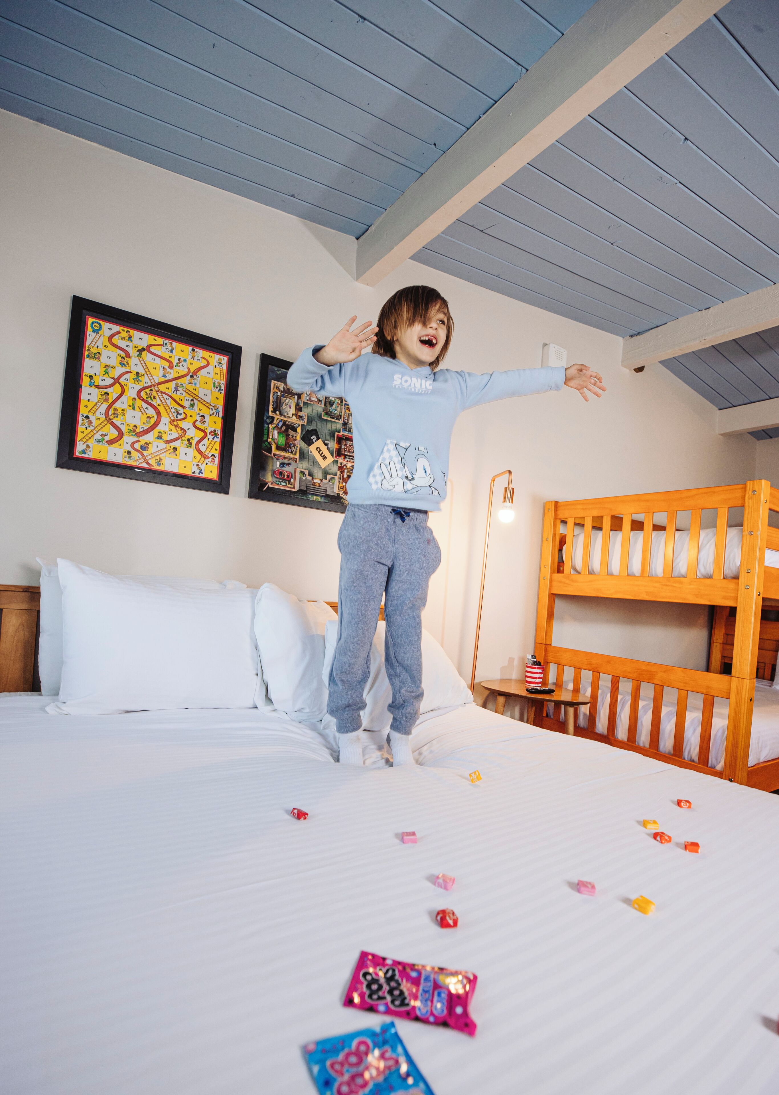 Un enfant saute sur un lit dans une chambre où se trouvent des lits superposés, un puzzle encadré et une horloge murale.