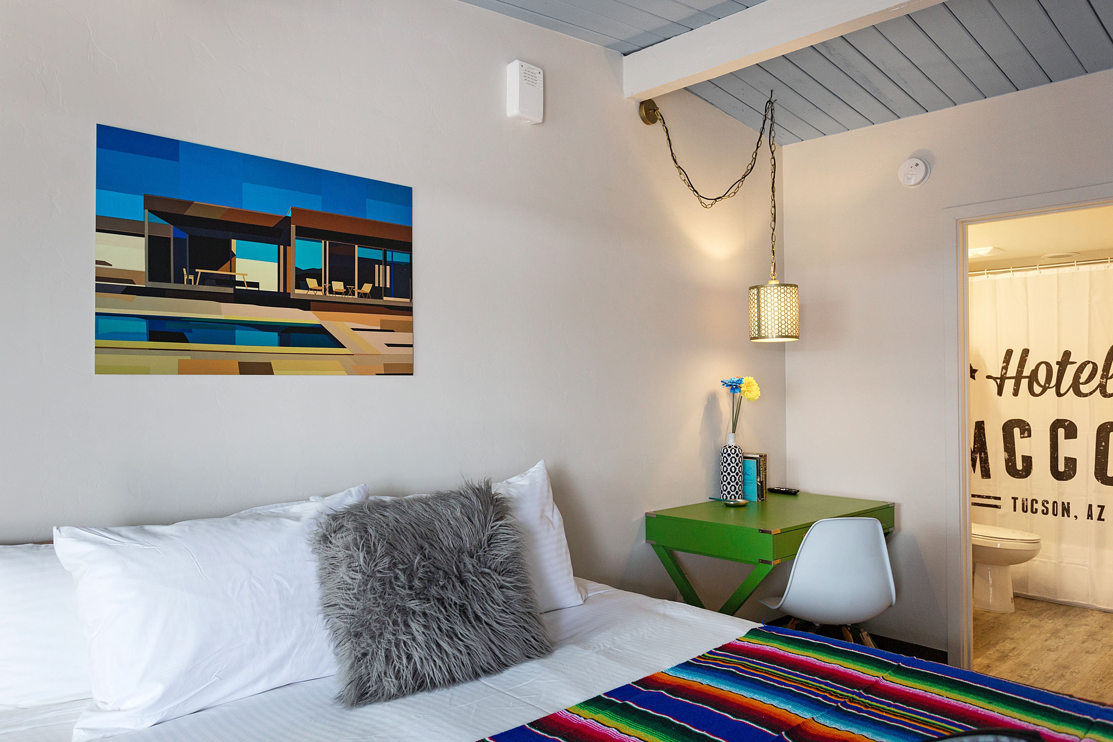 A hotel room with a bed, a desk, a chair, a painting of a building, and a sign reading "Hotel ACCO Tucson, AZ".