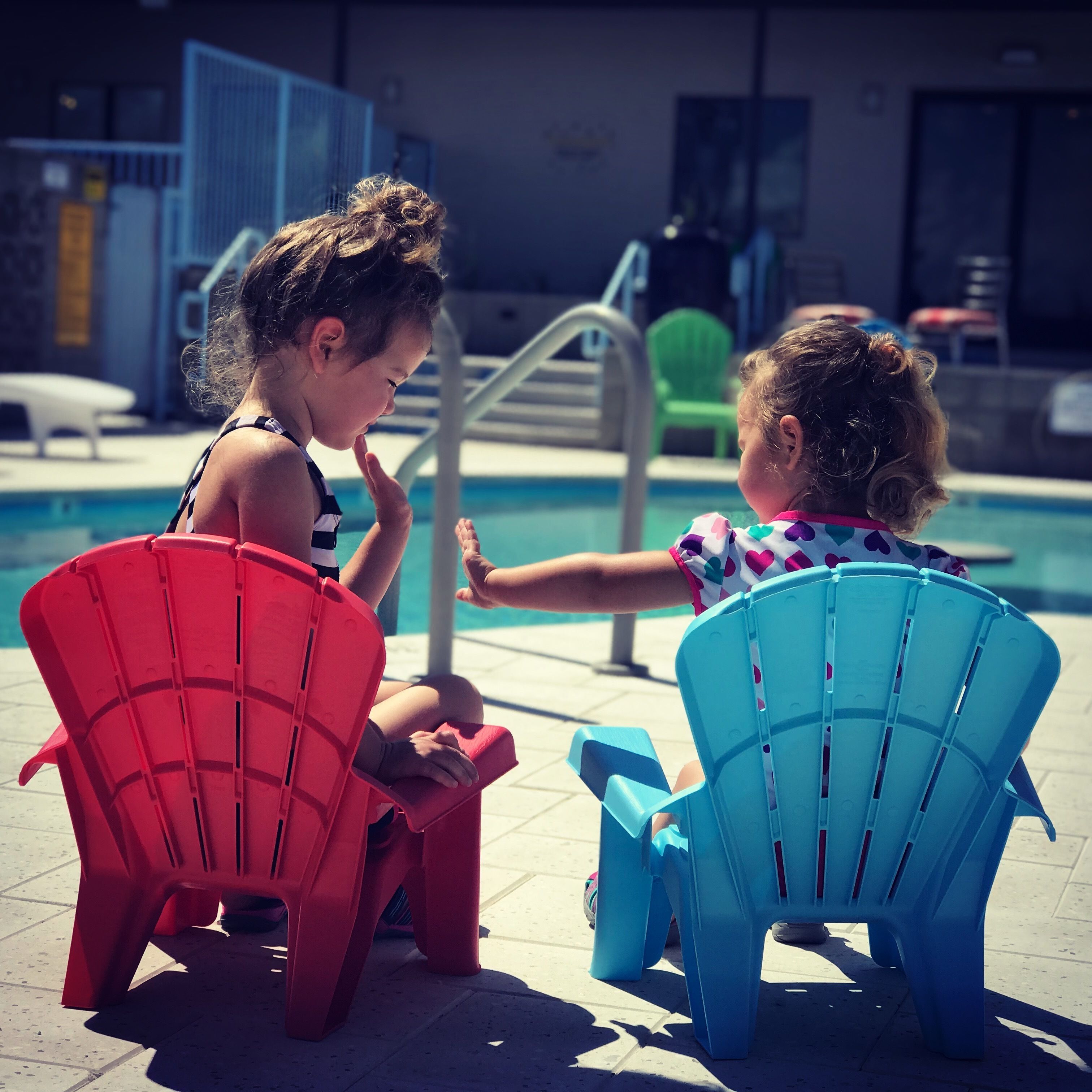 Two children sitting on chairs by a poolside.