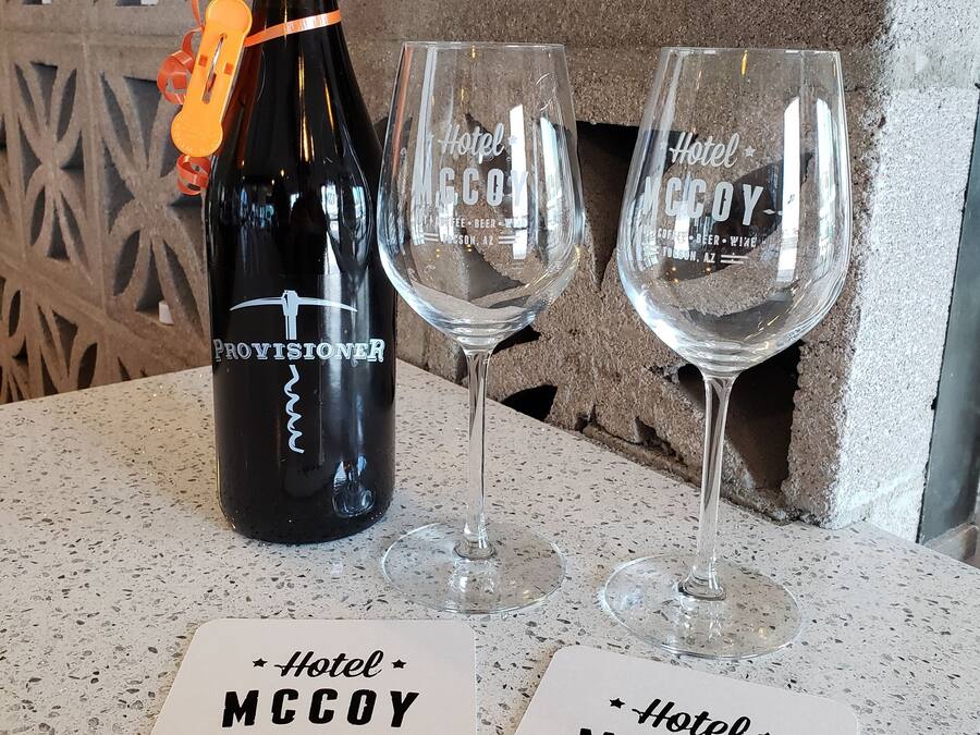 Een fles wijn, twee glaasjes en twee plaatsmatjes met de tekst 'Hotel McCoy' en 'Art, Coffee, Beer, Wine, Tucson, AZ' op een gekleurd aanrecht.