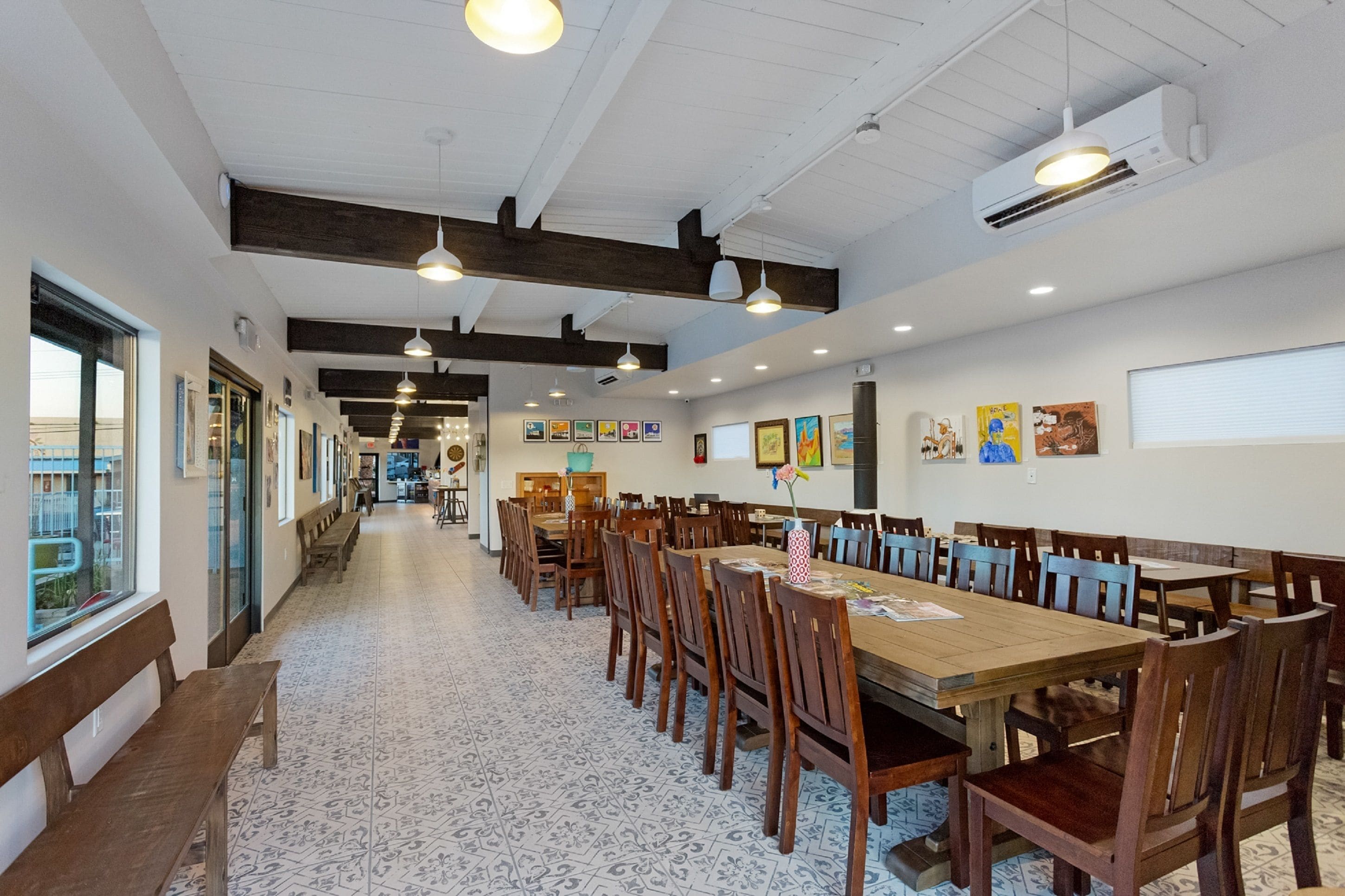 A spacious dining area with long wooden tables and chairs, white walls, and ceiling beams.