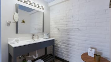 A modern bathroom with a white brick wall, a vanity with a mirror, two sinks, and a small table with a sign.