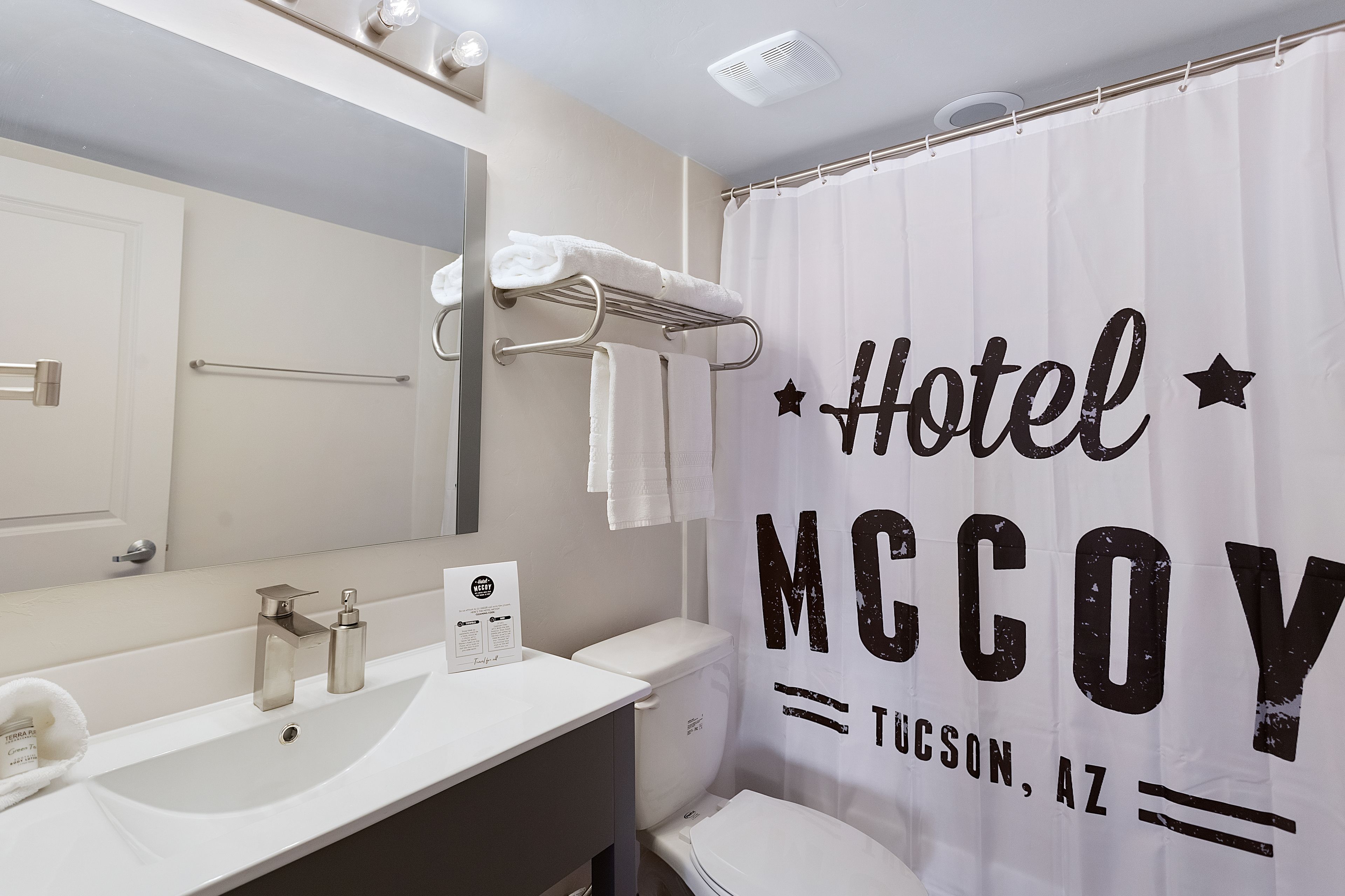 Un baño con lavamanos, espejo, toallero y cortina de ducha con la leyenda "Hotel McCoy Tucson, AZ".