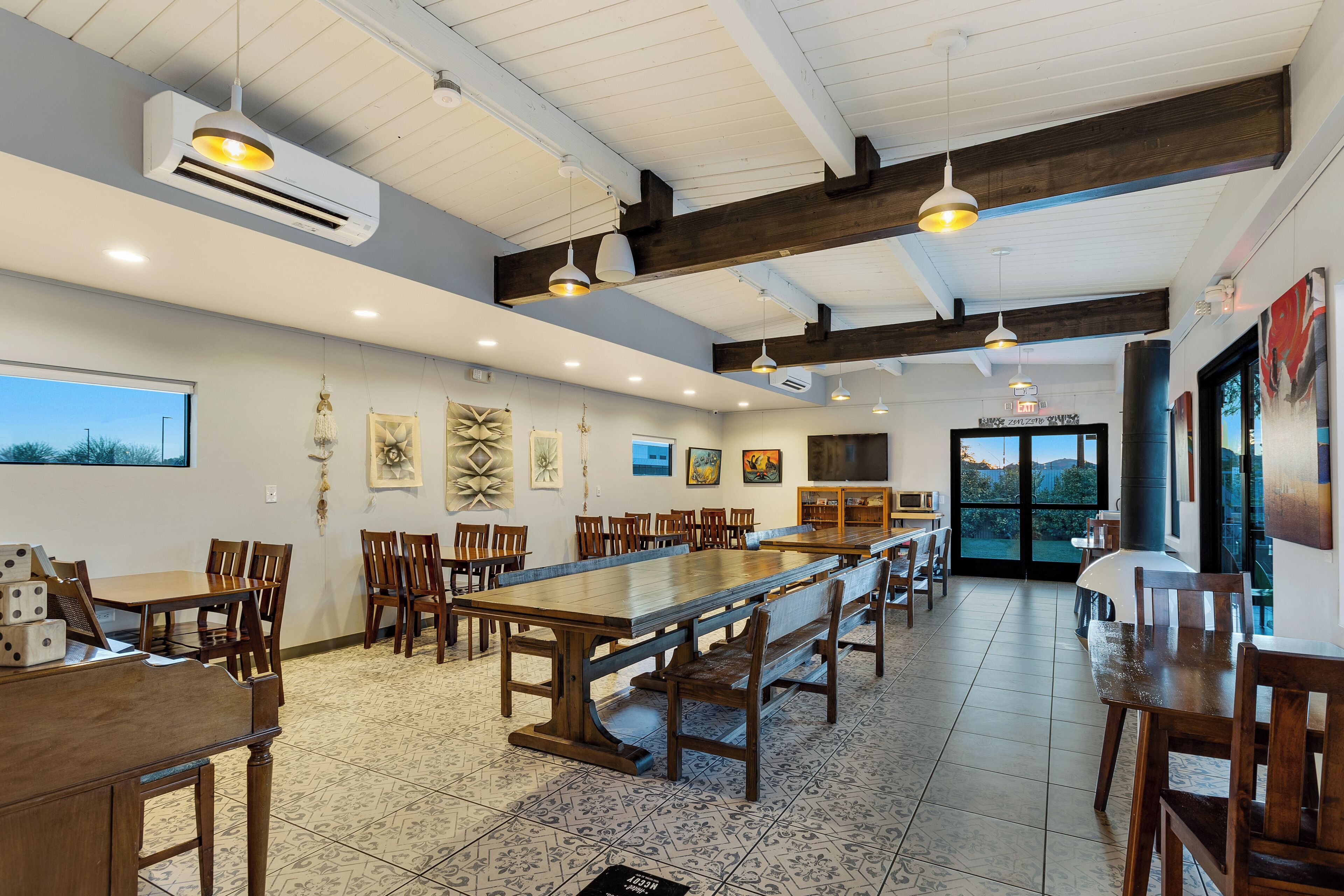 A spacious dining area with long wooden tables and chairs, a high ceiling with exposed beams, and a large window offering a view of the outdoors.