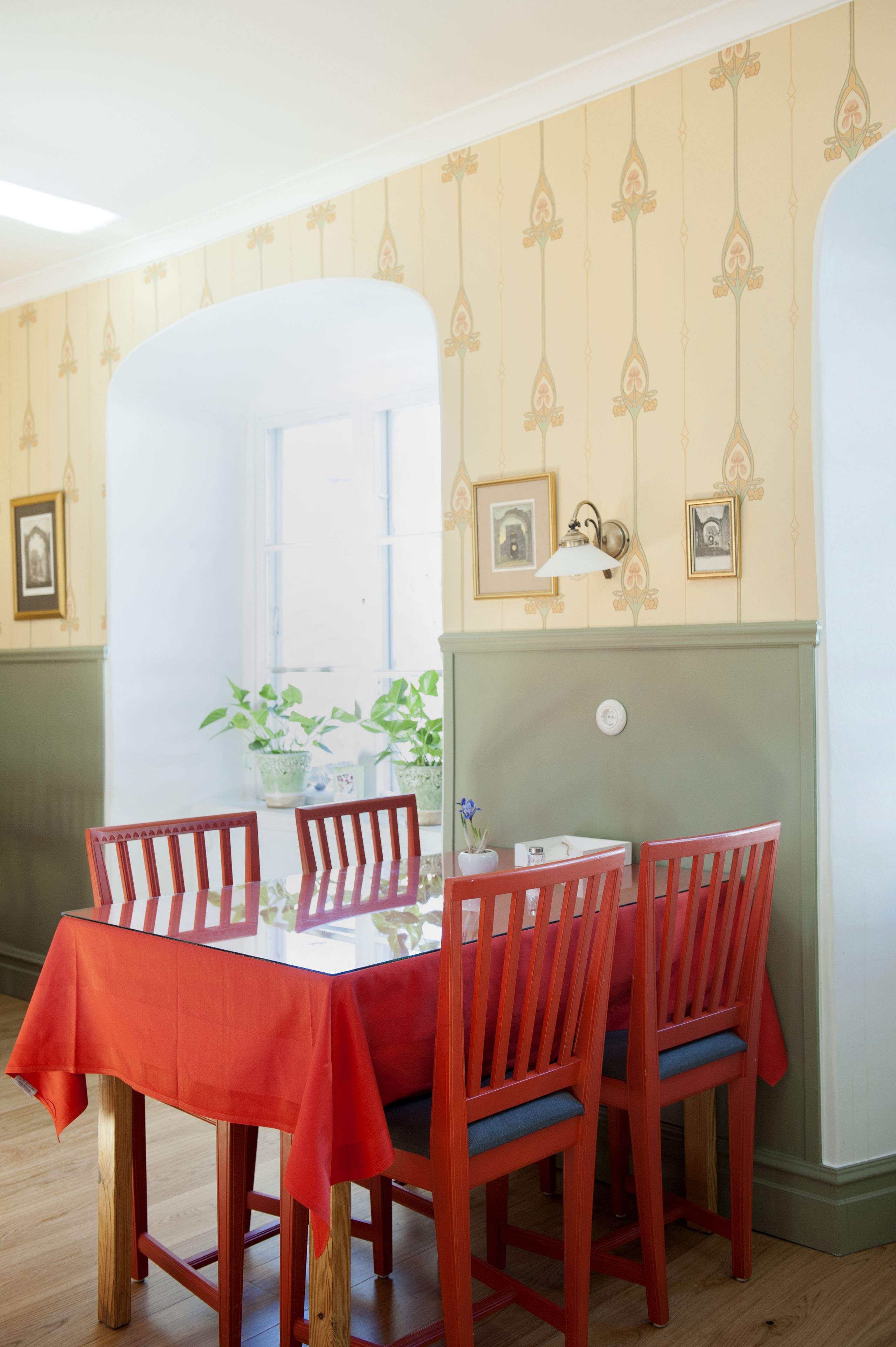 En matplats med ett bord dukat för fyra personer, röda stolar och en bordsduk. På fönsterbrädan står krukväxter och på väggen hänger inramade tavlor.