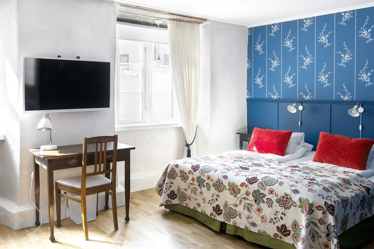 A bedroom with a bed, a desk, a chair, a TV, and a window with curtains.