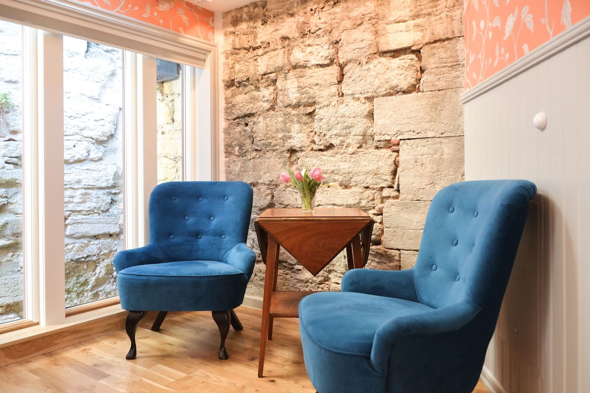 A room with two blue armchairs, a small wooden table with a vase of flowers, and a stone wall.