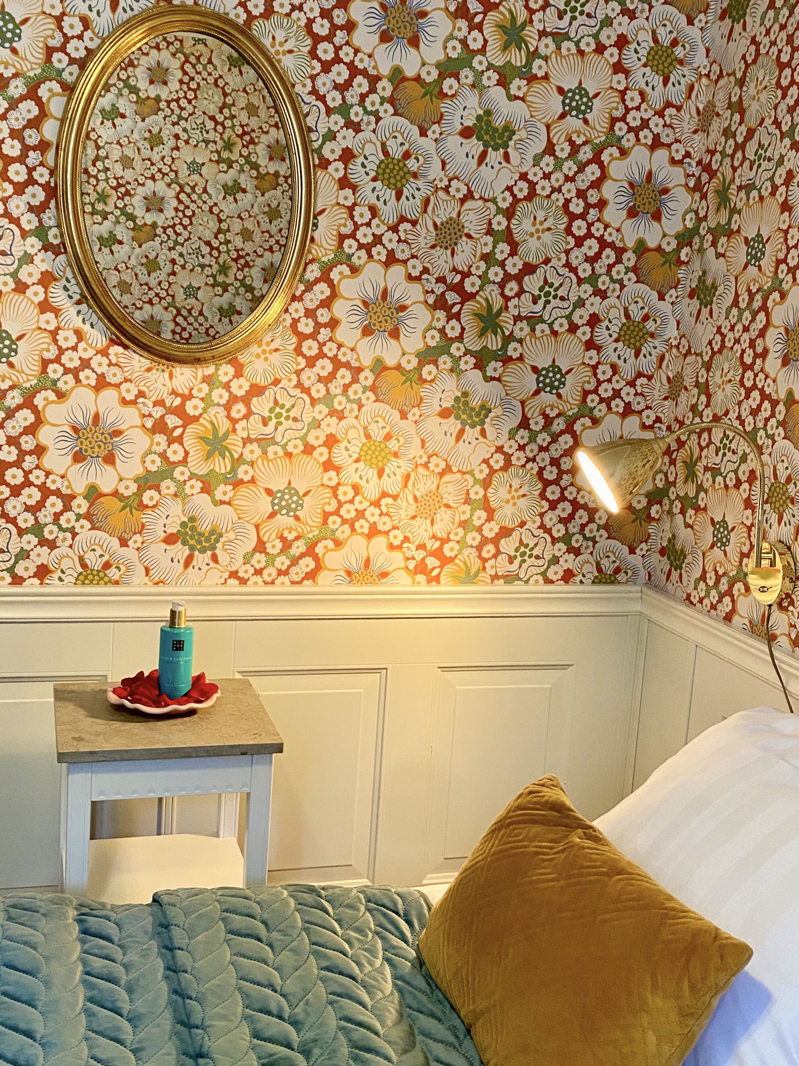 A room with floral wallpaper, a round mirror, a bedside table with a blue bottle, a gold lamp, and a bed with a blue quilt and a gold pillow.