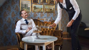 A man in a white shirt and tie sitting on a vintage chair in a room with ornate wallpaper and framed paintings on the wall. - Hotell St Clemens (Visby)