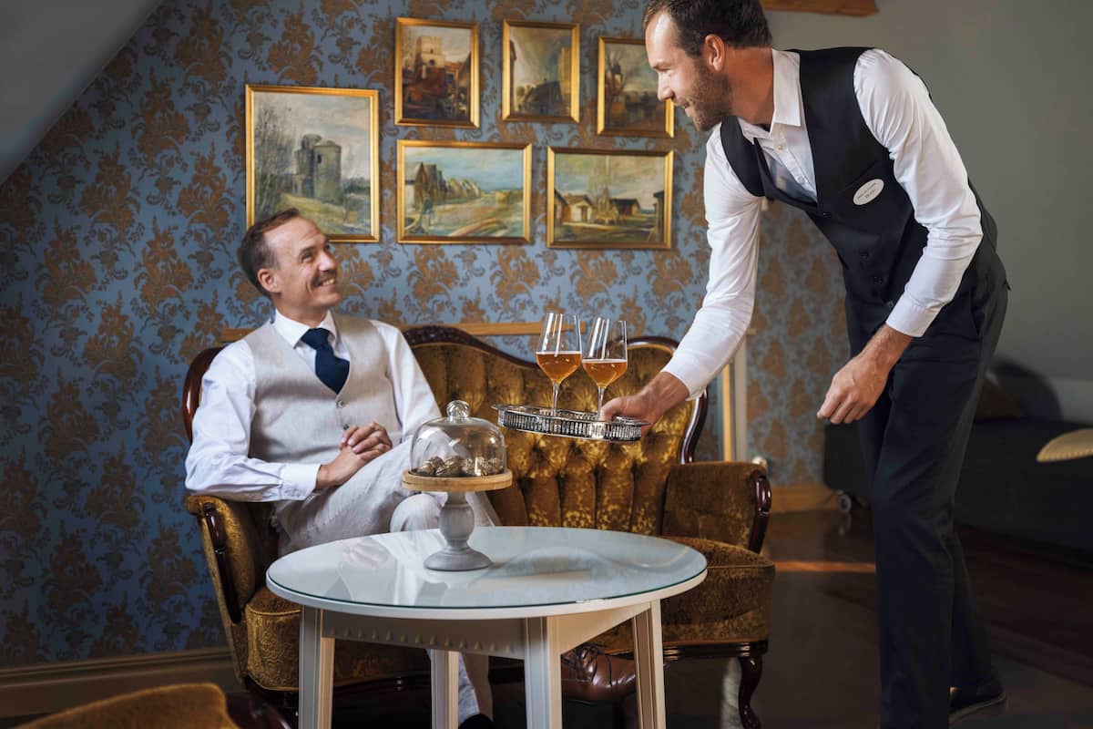 A man in a white shirt and tie sitting on a vintage chair in a room with ornate wallpaper and framed paintings on the wall.