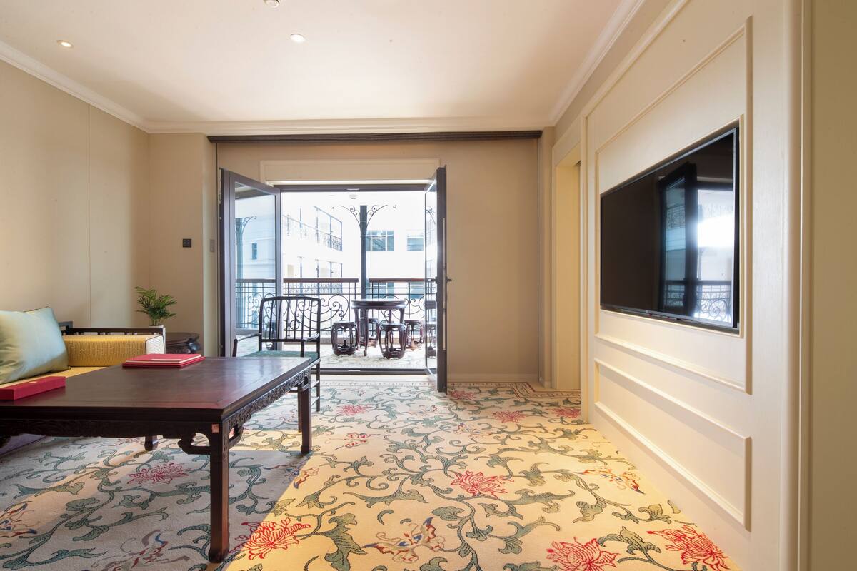 A hotel room with a large TV, a sofa, a dining table, and a patterned carpet.