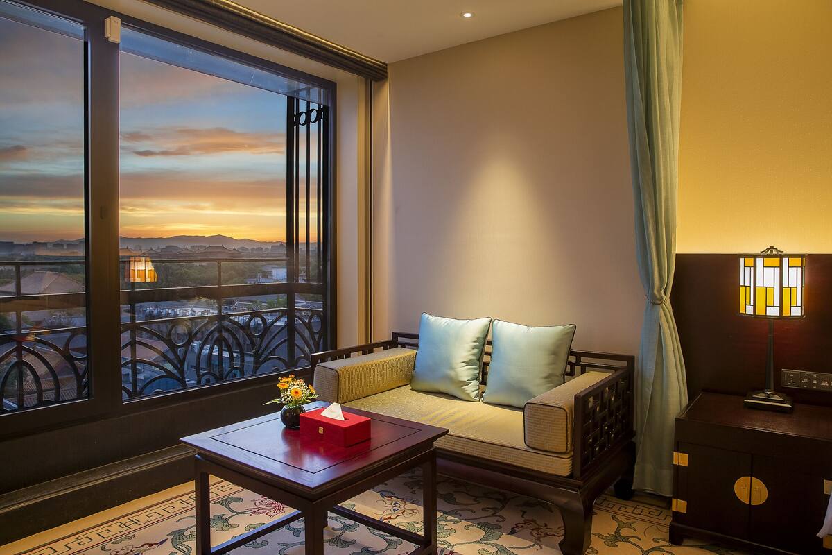 A hotel room with a large window offering a scenic view, a sofa with cushions, a small table with a tissue box, and a lamp.