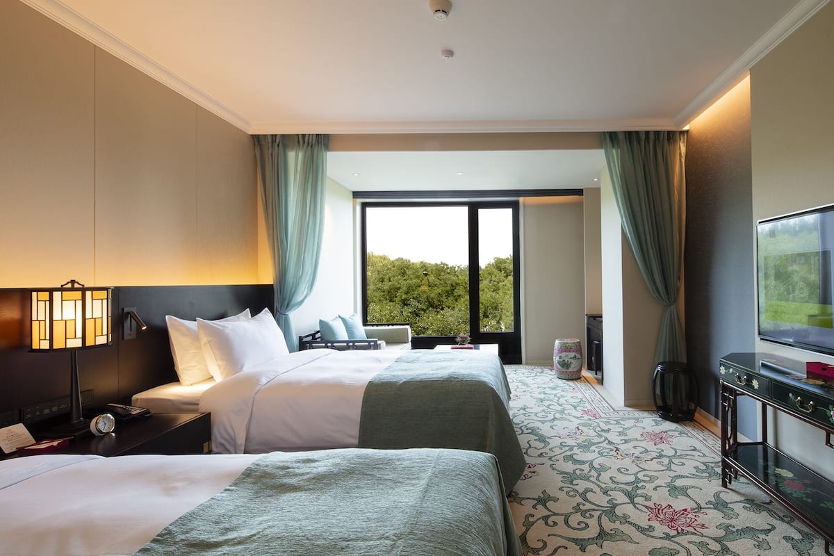 A hotel room with two beds, a TV, a desk, and a large window with curtains.