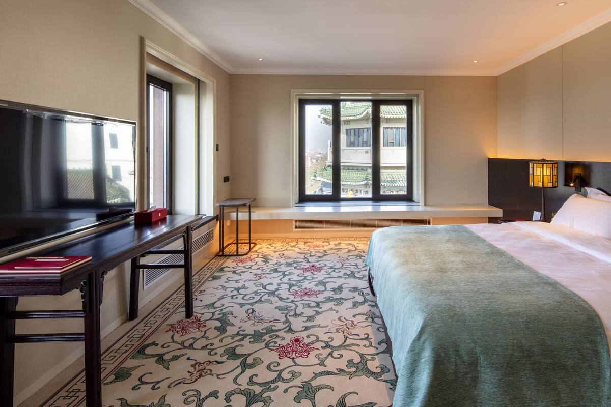 A hotel room with a bed, a desk with a TV, a window with a view of buildings, and a patterned carpet.