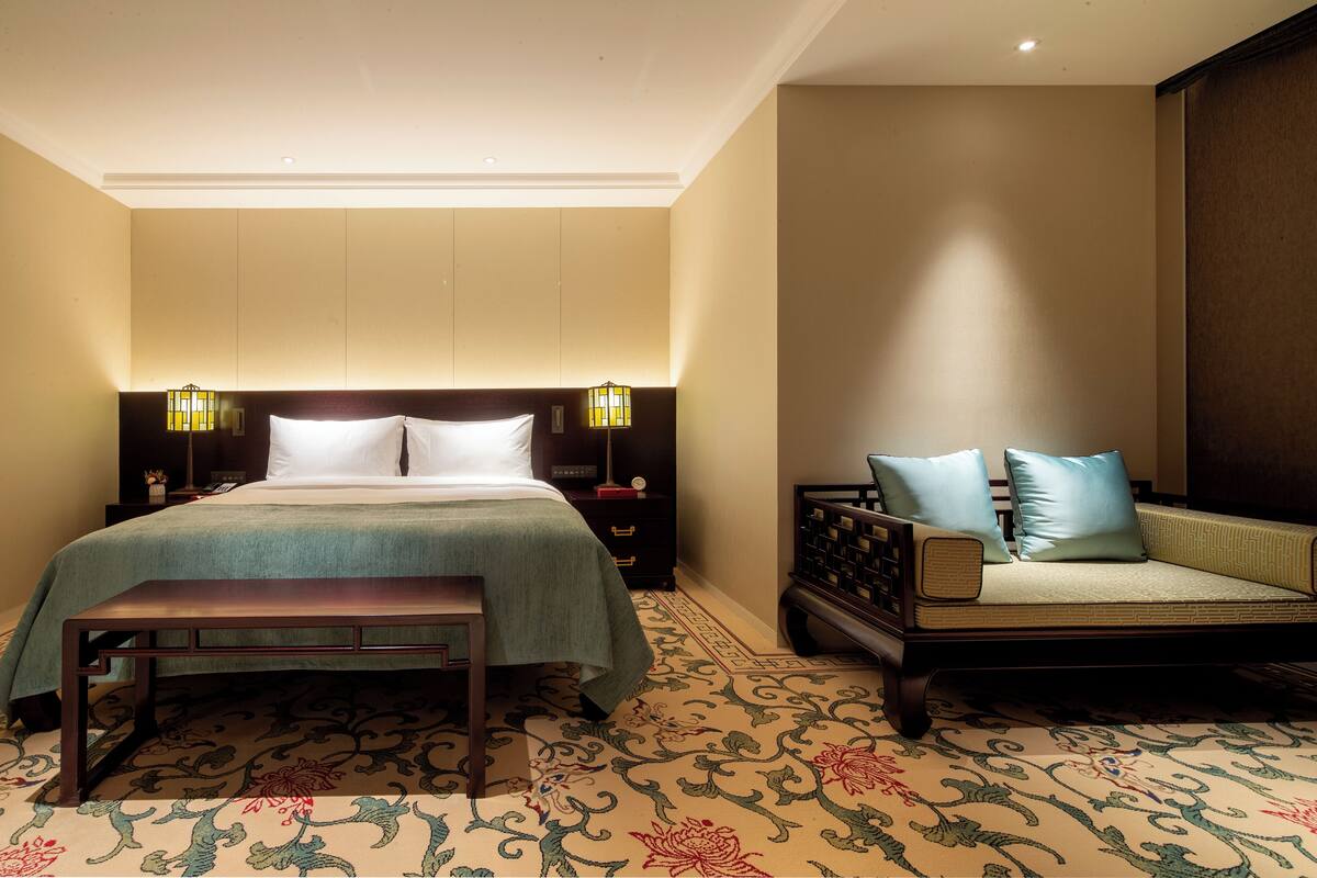 A hotel room with a bed, bedside tables, a bench, a sofa, and a patterned carpet.